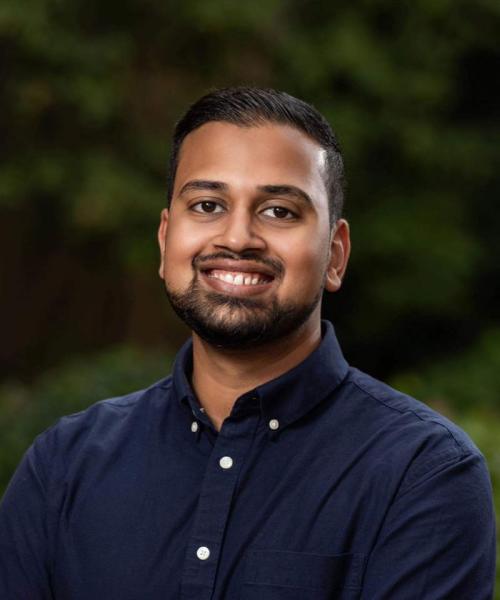 Male with brown skin and beard who is smiling. Individual is wearing blue button up t-shirt, there is a greenery background. 