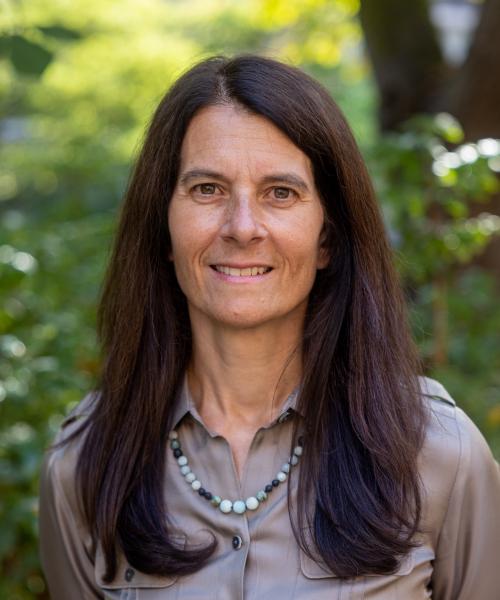 Picture of Jennifer Hoffman taken outside Miller Hall with green leafy background. Jennifer is smilling, has long brown hair and is wearing a brown blouse & light green necklace. 