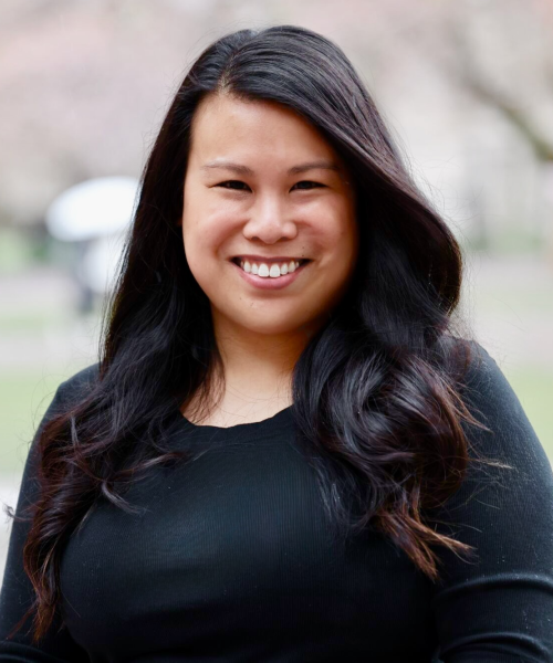A woman with long, dark hair is smiling in front of cherry blossom trees