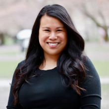 A woman with long, dark hair is smiling in front of cherry blossom trees