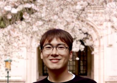 A smiling person with glasses stands in front of a building, surrounded by blooming cherry blossoms, creating a serene and joyful spring atmosphere.